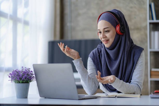 Hijab-wearing female Muslim students study online with laptop and headphones in remote online meetings.
